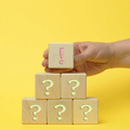 A hand places a small wooden block with an exclamation mark on top of a pyramid of wooden blocks marked with question marks, set against a bright yellow background, symbolizing clarity or answers emerging from uncertainty.